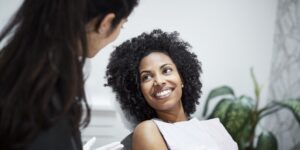 Smiling woman with curly hair at a dental clinic, interacting with a friendly dentist or healthcare professional during a checkup, in a modern medical office setting.