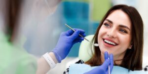 A smiling woman sitting in a dental chair during a routine cleaning, with a dental professional wearing gloves holding tools near her mouth.
