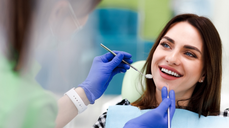 A smiling woman sitting in a dental chair during a routine cleaning, with a dental professional wearing gloves holding tools near her mouth.